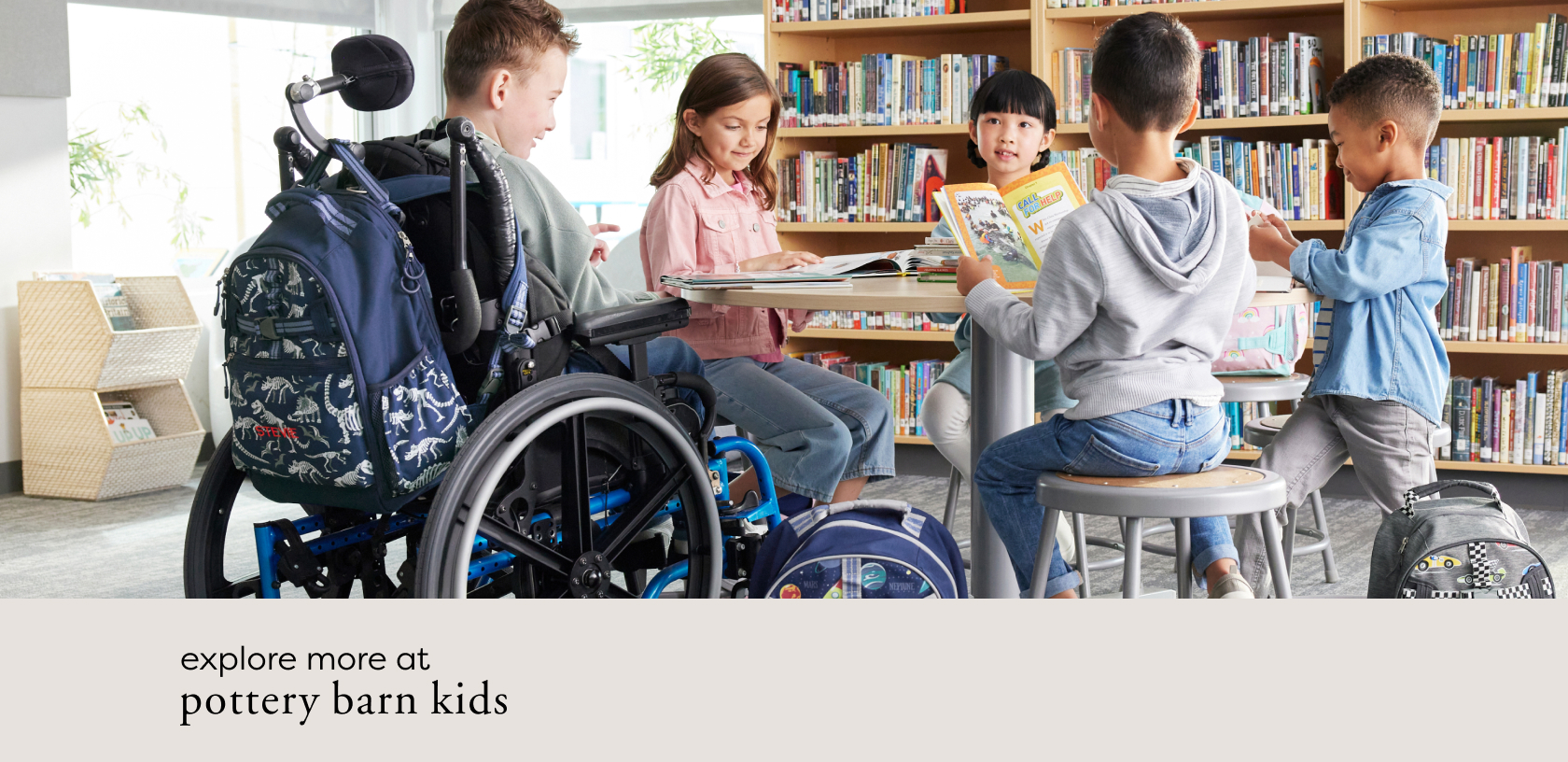 Image shows a group of five school-aged children sitting at a table in a school library. Some are reading books and some are talking to each other. One boy in the group is sitting in a wheelchair with a Pottery Barn Kids Mackenzie Adaptive Backpack in Dino Bones print attached to his chair.