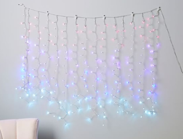 Strings of fairy lights waterfall down a wall in an ombre pattern from pink to purple to blue to teal. 