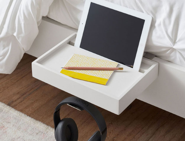 A bedside shelf next to a bed with a tablet, pad of paper, and headphones attached to it.