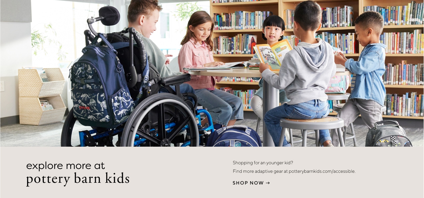 Image shows a group of five school-aged children sitting at a table in a school library. Some are reading books and some are talking to each other. One boy in the group is sitting in a wheelchair with a Pottery Barn Kids Mackenzie Adaptive Backpack in Dino Bones print attached to his chair.