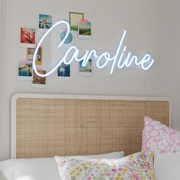 A custom personalized neon LED sign showing the name ’Caroline’ hangs above a rattan dorm headboard