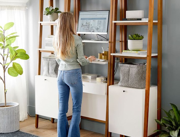 Woman standing in front of West elm modern adjustable wall desk narrow bookshelf using computer.