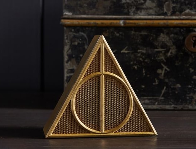 A Bluetooth speaker shaped like the Deathly Hallows symbol from Harry Potter sits on a wooden tabletop.