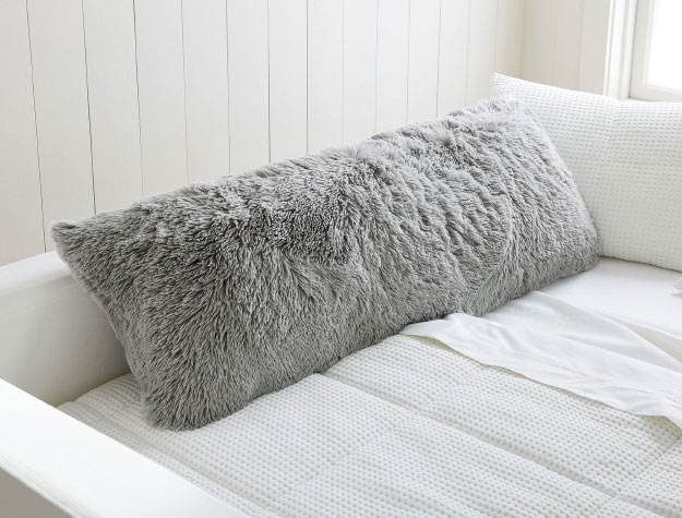 A grey fluffy body pillow on top of a bed with white sheets.