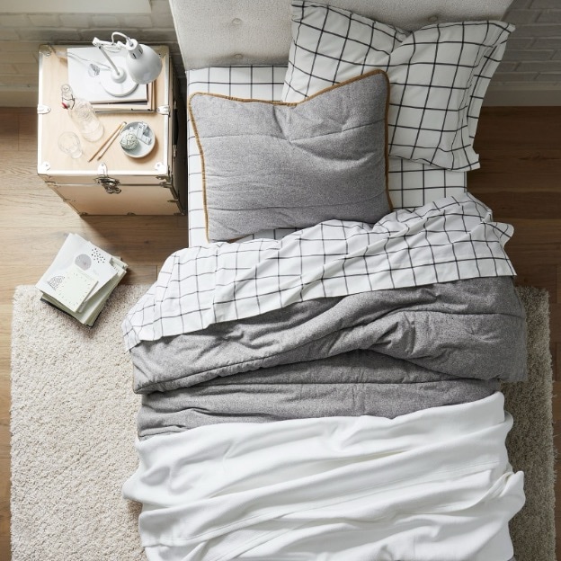 A cozy dorm bed made up with pillows, plaid twin XL sheets, a gray reversible comforter, and a white throw blanket on top.