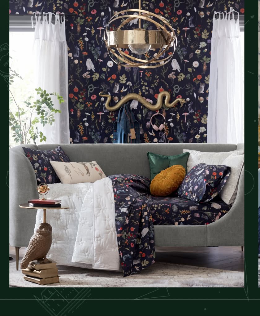 Harry Potter sample designed room. Shows a couch on top of a faux fur rug. The couch is draped with a Harry Potter theme plants and potions blanket in navy with gold, red, and green accents and iconography. The wallpaper behind the couch matches the Harry Potter themed blanket.