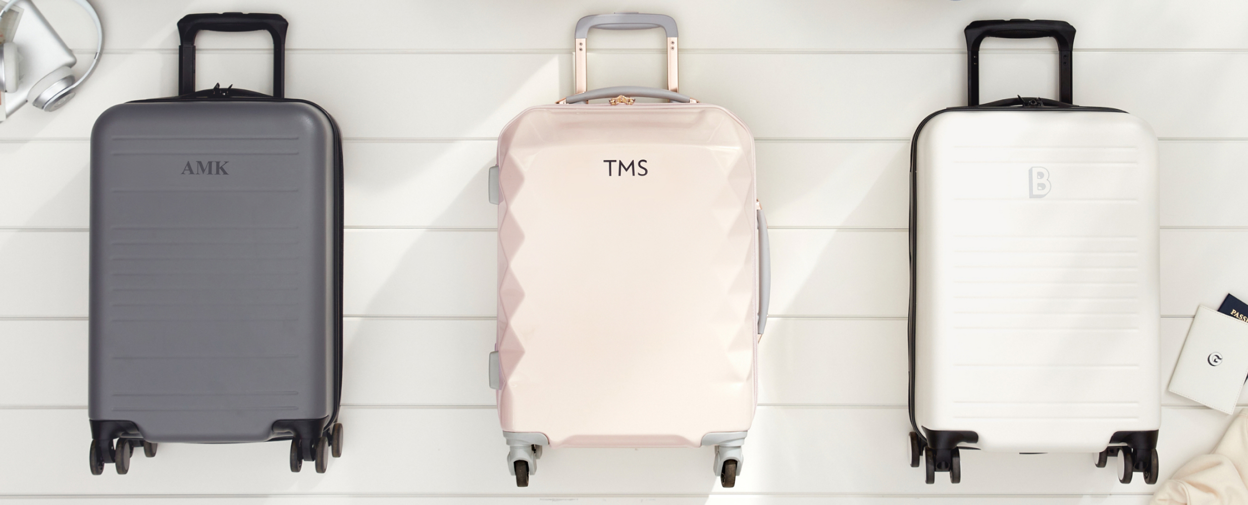 3 Luggage bags with similar color schemes including dark gray, light pink, and white, all of which have monogrammed personalizations, lie on top of an off white wood paneled background.
