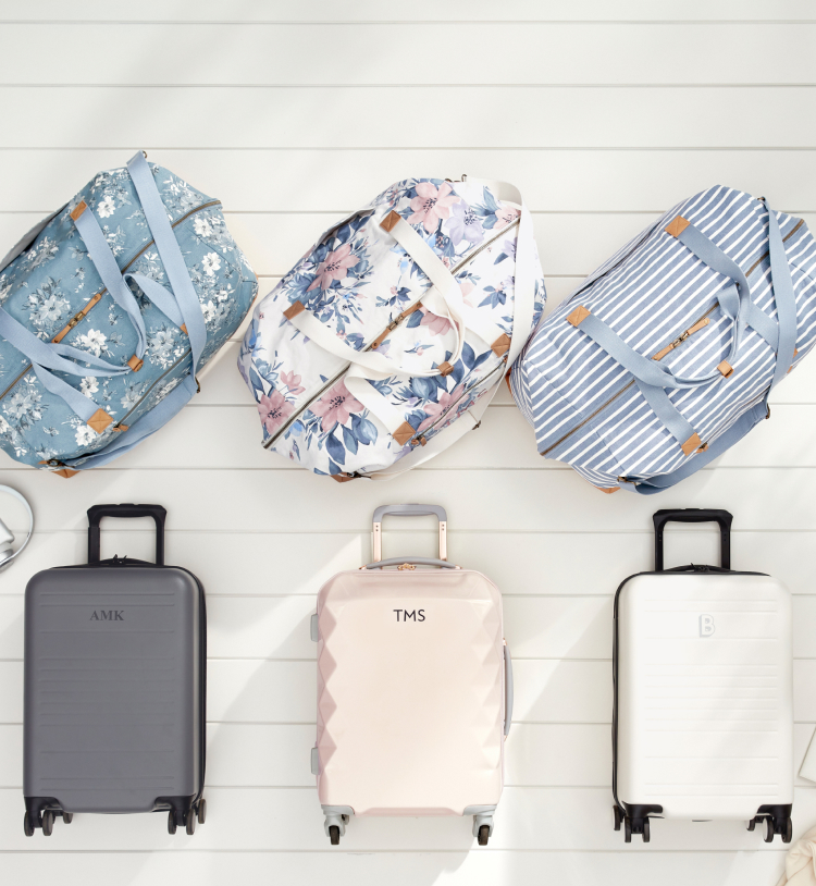3 duffel bags with flower and striped patterns in blue and white colors, are stacked in a row. 3 Luggage bags with similar color schemes including dark gray, light pink, and white, all of which have monogrammed personalizations, lie in a row beneath the duffel bags. All of these luggage options lie on top of an off white wood paneled background.