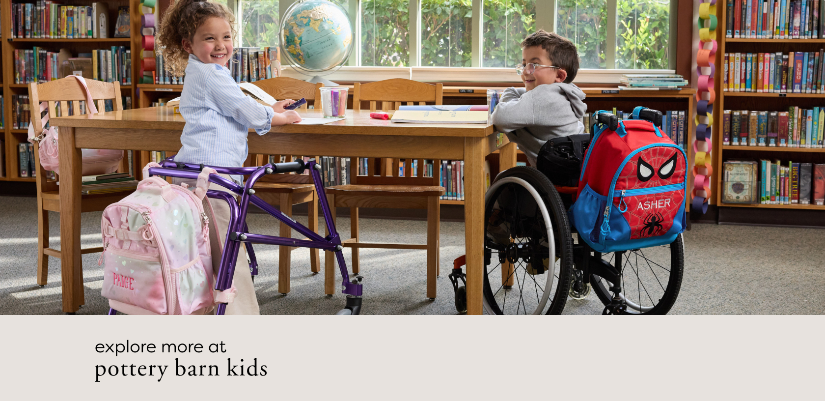Image shows a pair of school-aged children at a table in a school library. On the left, a girl in a light-blue striped shirt is turned inwards facing the camera and smiling. She stands in front of a purple accessibility walker. The walker has an adaptive backpack in pink heart print, personalized with the name 'Paige' on it. On the right, a boy is sitting in a wheelchair turned inwards facing the camera and smiling. An adaptive backpack in Spider-Man™ print, personalized with the name 'Asher', is attached to his chair.