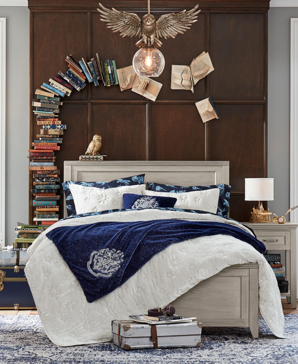 Harry Potter sample designed room. Shows a bedroom featuring a Hogwarts insignia themed velvet quilt sham in navy blue. It contrasts against the dark brown wooden bedframe with a golden statue of an owl above the headboard. Books are stacked to the side of the bed, and create a small arch over the headboard, seemingly magically.