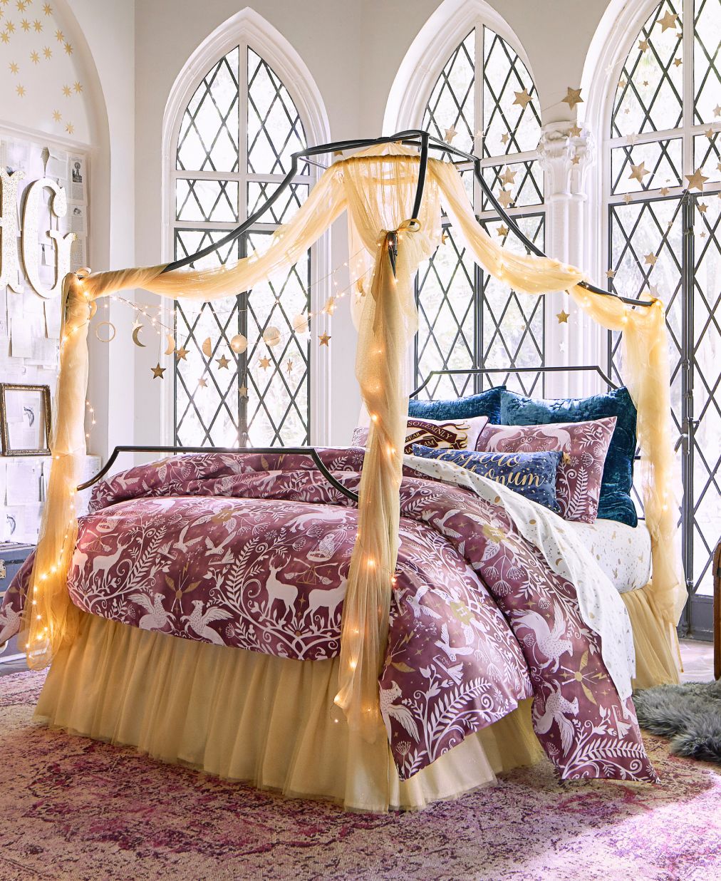 Harry Potter sample designed room. Shows a bedroom featuring the wizarding world enchanted night sky sheet set in a mauve color. Gold accents adorn the room and small warm lights adorn the the frame of the bed. The room has tall slender windows with lattice work adorning the outside. Soft lighting illuminates from outside.