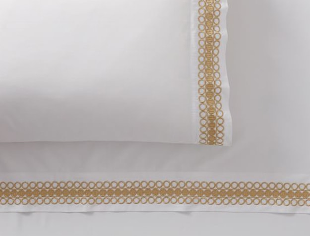 A white bedspread with gold embroidery on the pillowcase and sheet edges rests on a bed.