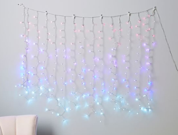 Strings of fairy lights waterfall down a wall in an ombre pattern from pink to purple to blue to teal. 