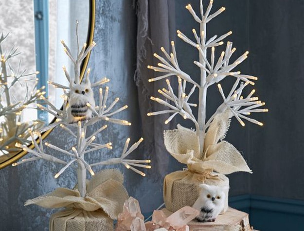 Two white mini trees with twinkle lights sit on top of a table.
