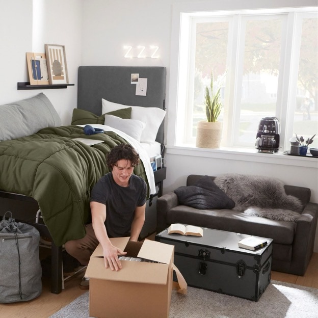A teenage guy opens a box next to a storage trunk and sleeper sofa in his dorm room.