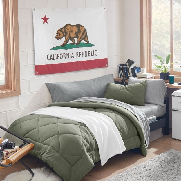 This dorm bed features olive green and grey twin XL bedding, a dark grey body pillow, and a California state flag above