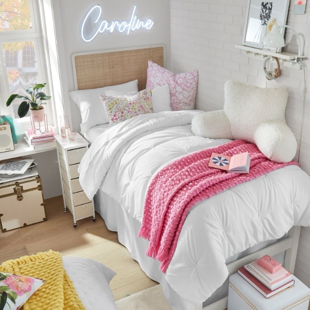 This dorm bed features a twin XL microfiber pintuck comforter in white along with dceorative pillows and a pink throw blanket