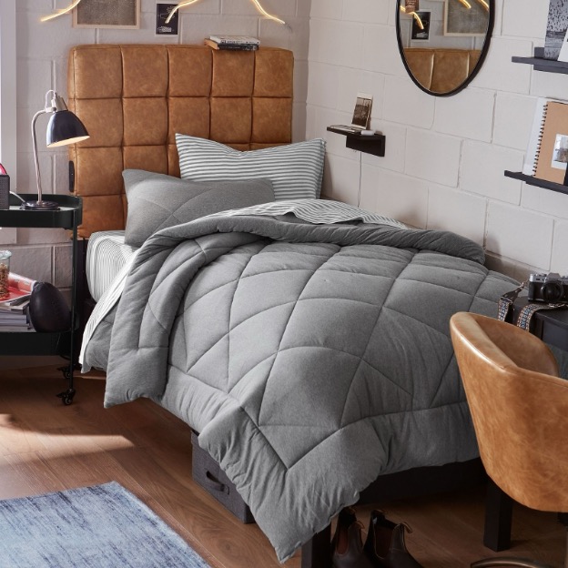 A guy’s dorm room features gray twin XL bedding, striped sheets, and a faux leather tech no-nails headboard.