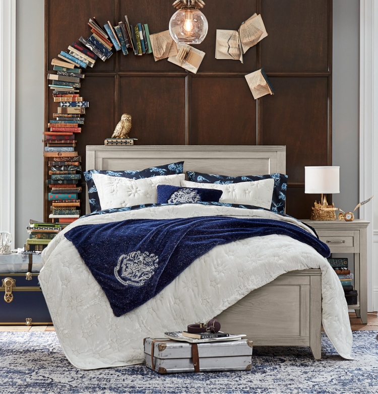 Harry Potter sample designed room. Shows a bedroom featuring a Hogwarts insignia themed velvet quilt sham in navy blue. It contrasts against the dark brown wooden bedframe with a golden statue of an owl above the headboard. Books are stacked to the side of the bed, and create a small arch over the headboard, seemingly magically.