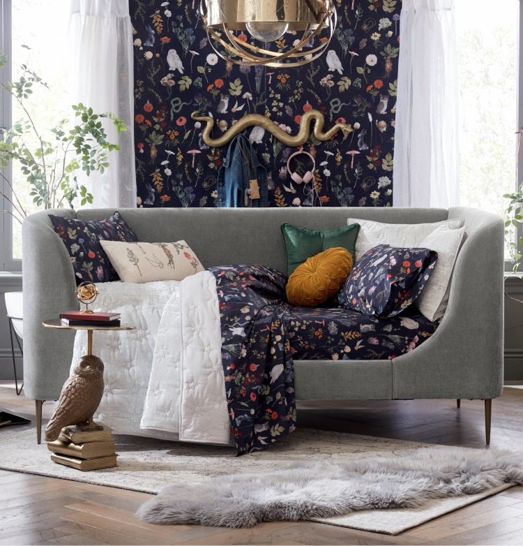 Harry Potter sample designed room. Shows a couch on top of a faux fur rug. The couch is draped with a Harry Potter theme plants and potions blanket in navy with gold, red, and green accents and iconography. The wallpaper behind the couch matches the Harry Potter themed blanket.