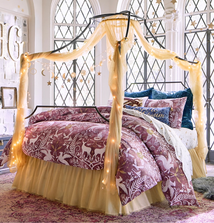 Harry Potter sample designed room. Shows a bedroom featuring the wizarding world enchanted night sky sheet set in a mauve color. Gold accents adorn the room and small warm lights adorn the the frame of the bed. The room has tall slender windows with lattice work adorning the outside. Soft lighting illuminates from outside.