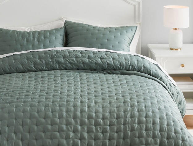 A sage quilt with square stitching lays on top of a bed.