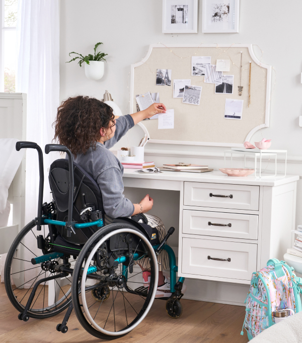 Image shows a teenage girl in a wheelchair sitting at a Pottery Barn Teen Hampton Desk - ADA in her bedroom. She is reaching above the desk to place a note onto a pinboard on the wall.